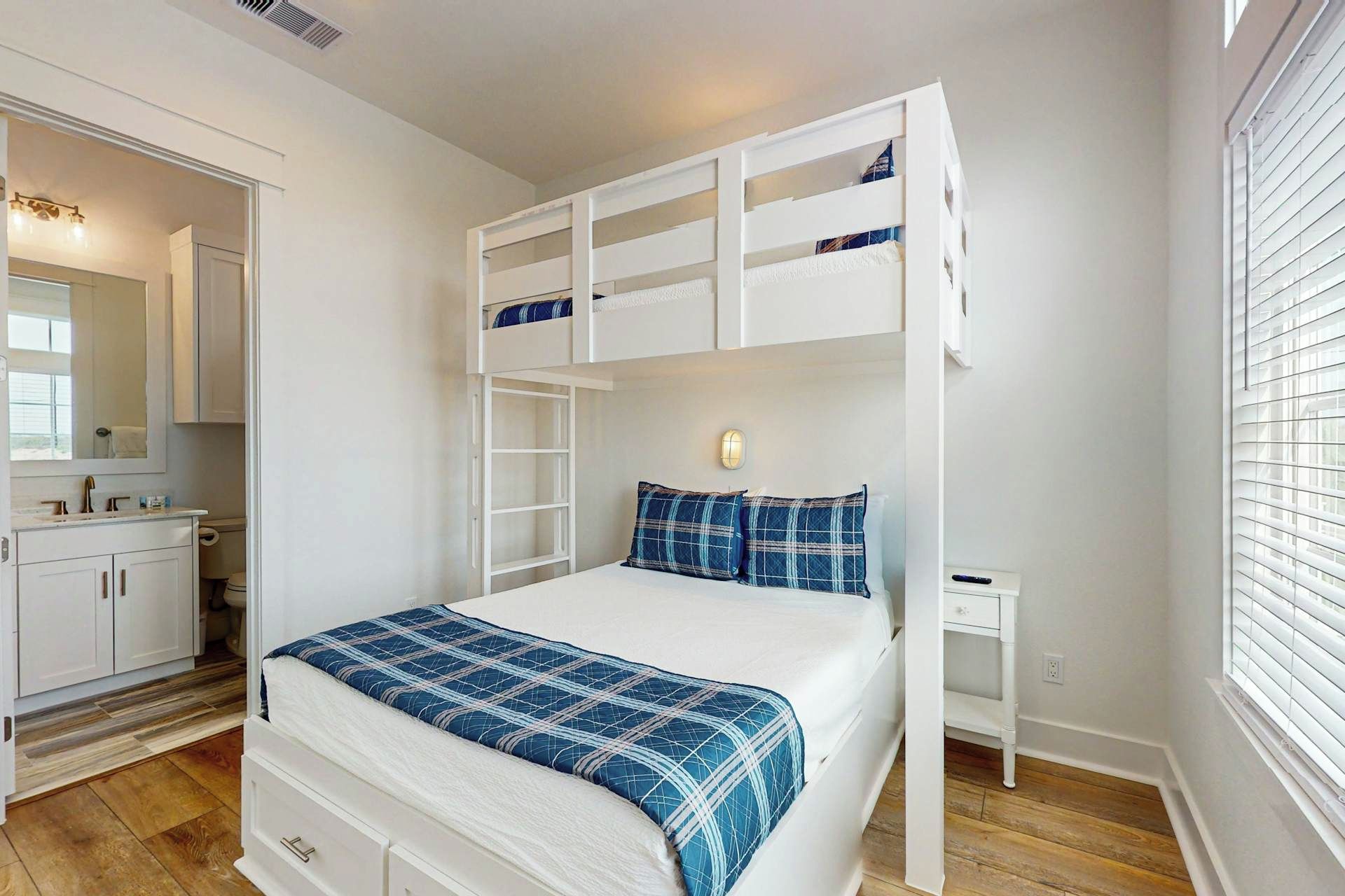 A bedroom with a bunk bed, a bedside table, a nightstand, a mirror, and a window with blinds.