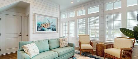 A spacious living room with a teal sofa, wicker chairs, a round coffee table, and a ceiling fan.