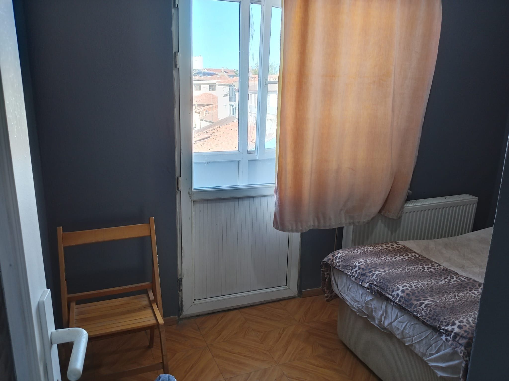 Una habitación pequeña con una cama individual, una silla, un radiador y una ventana con cortina.