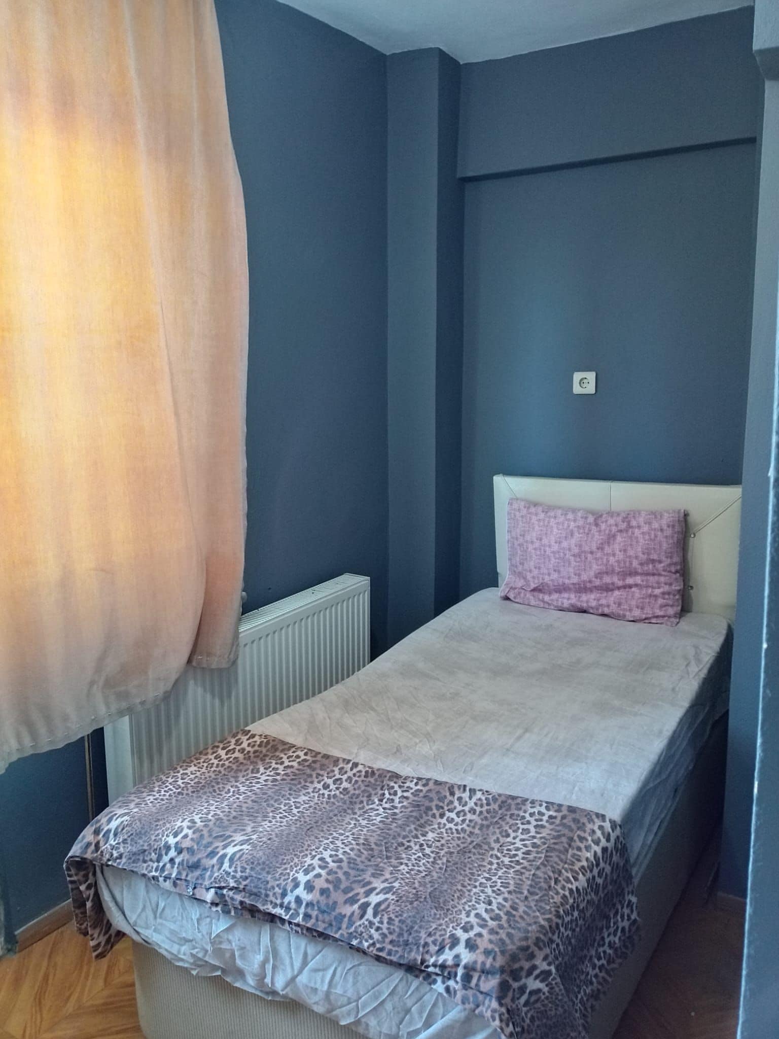 Una habitación pequeña con una cama individual, cabecera sencilla, un radiador y un enchufe en la pared.