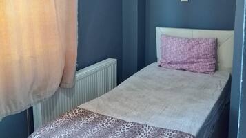 Una habitación pequeña con una cama individual, cabecera sencilla, un radiador y un enchufe en la pared.