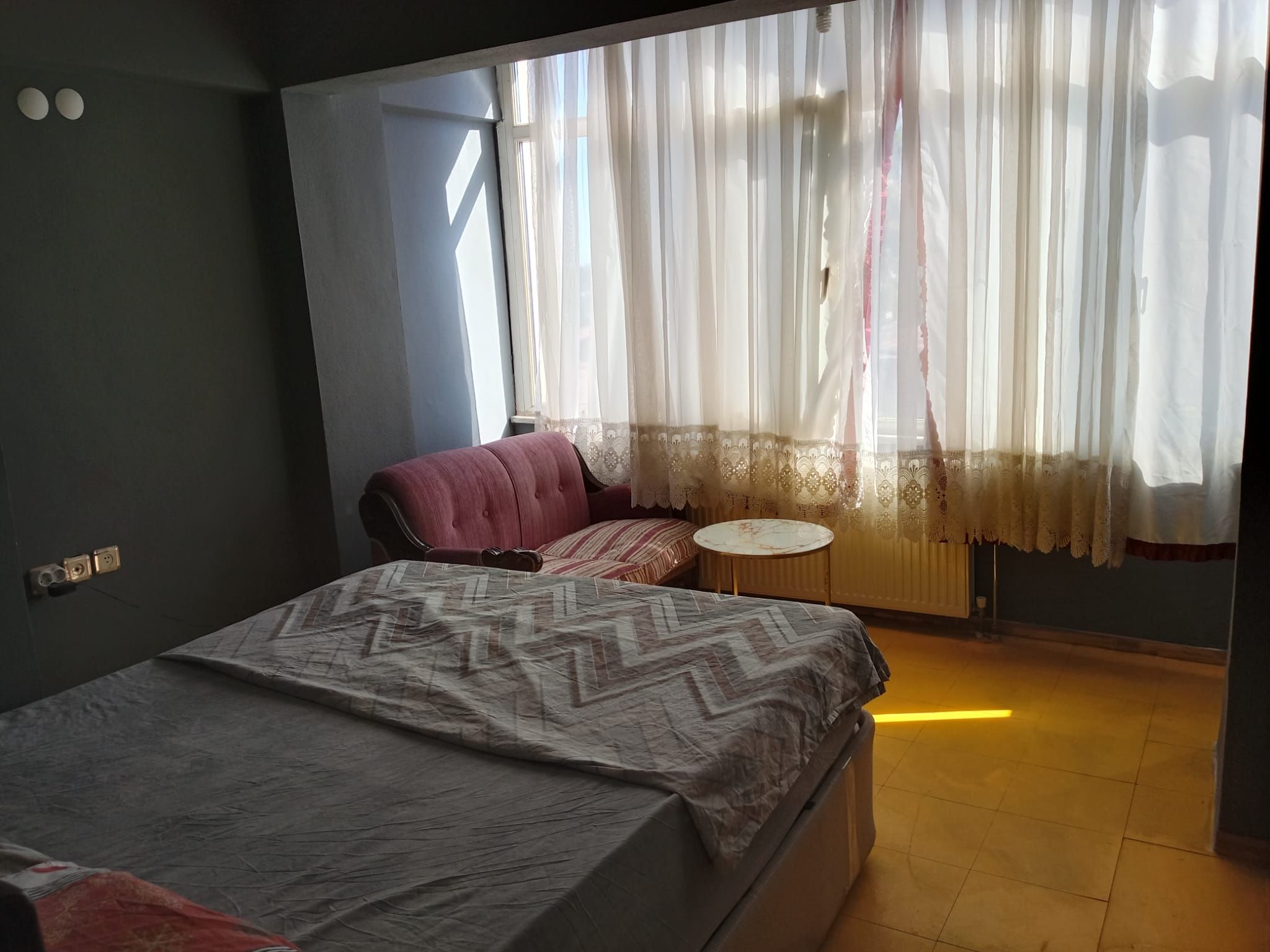 Un dormitorio con una cama, una silla, una mesita y una ventana con cortinas translúcidas.