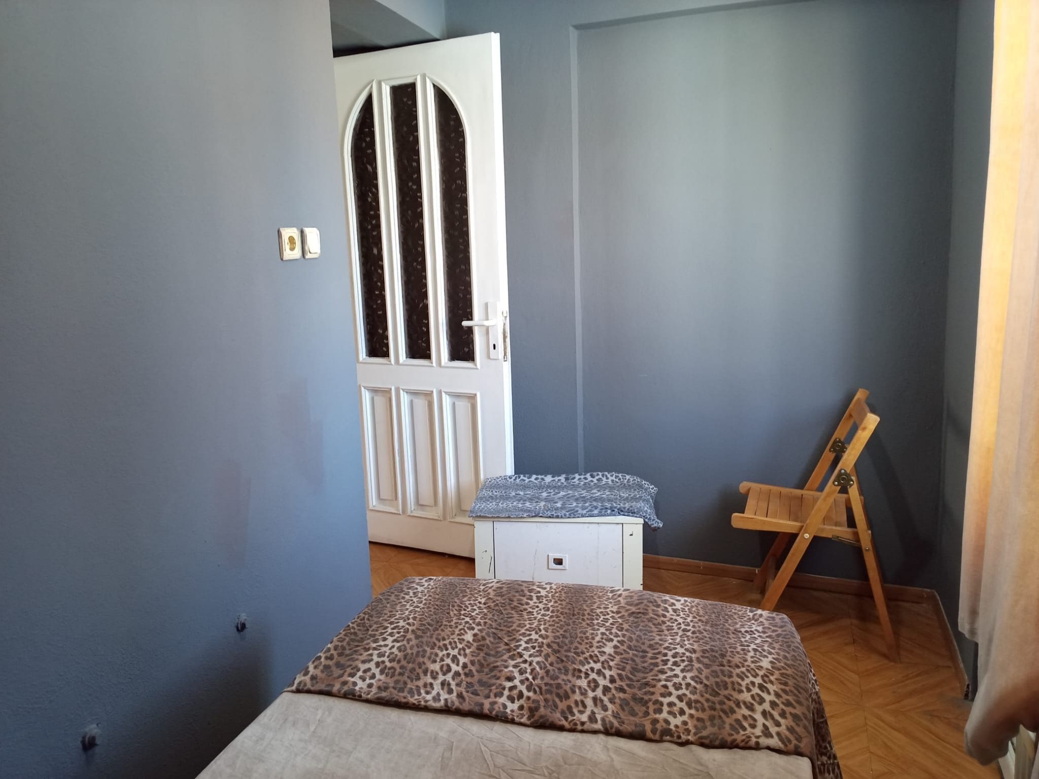 A bedroom with a bed, a small white dresser, a wooden easel, and a door with a window.