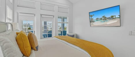 Een slaapkamer met een bed, een raam met zonwering, een schilderij van een strand en uitzicht op buiten.