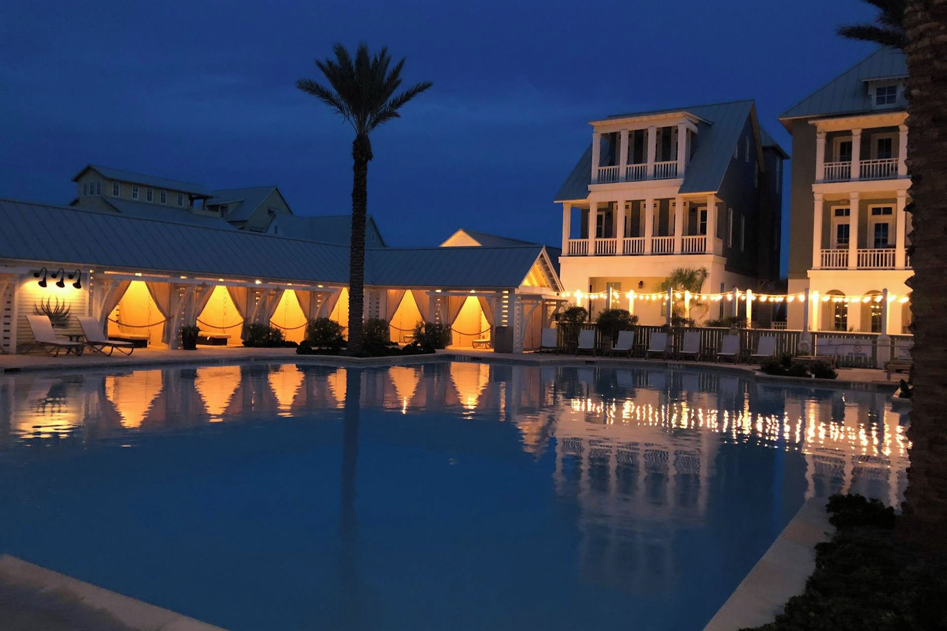 A pool area with a reflection of a building and palm trees, surrounded by a covered seating area with string lights.