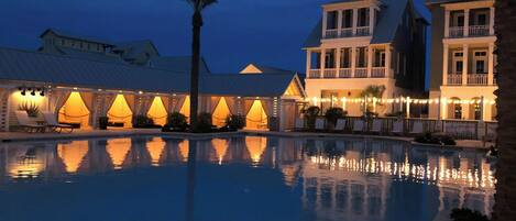 A pool area with a reflection of a building and palm trees, surrounded by a covered seating area with string lights.