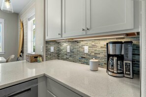A kitchen with white cabinets, a tiled backsplash, a coffee maker, and a surfboard. - Soñador | Golf Views | Steps to Beach | Game Room (Port Aransas)