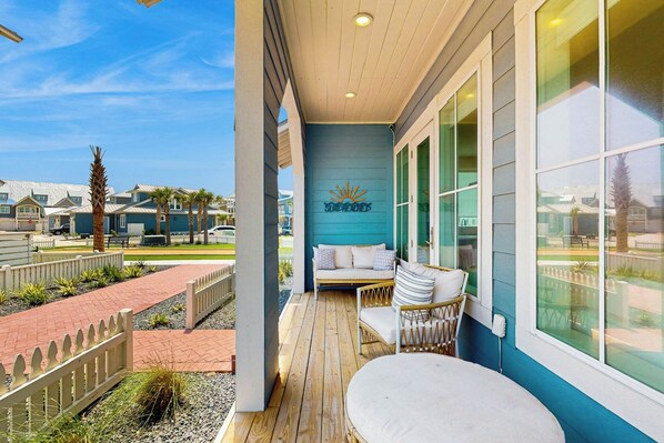 A covered outdoor seating area with a wooden deck, a white cushioned sofa, and a wicker chair. - Sun-Drenched On Sundew | Steps to Beach | Resort (Port Aransas)