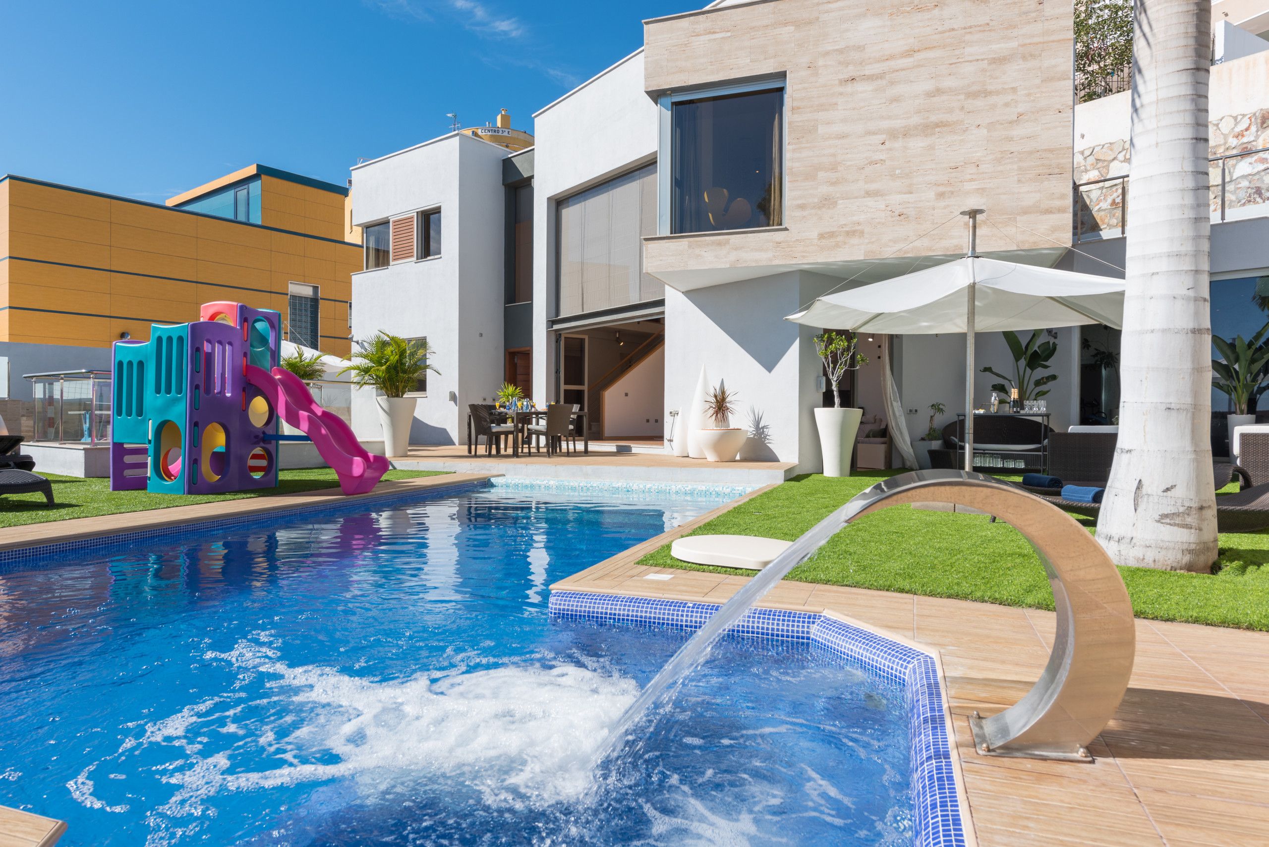 A swimming pool with a slide, a patio area with outdoor furniture, and a modern building with large windows.
