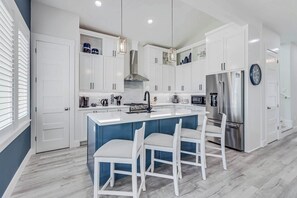A modern kitchen with white cabinetry, a central island with bar stools, a stainless steel refrigerator, and a built-in oven. - Sunrise Villa | Steps to Beach | Resort Amenities (Port Aransas)