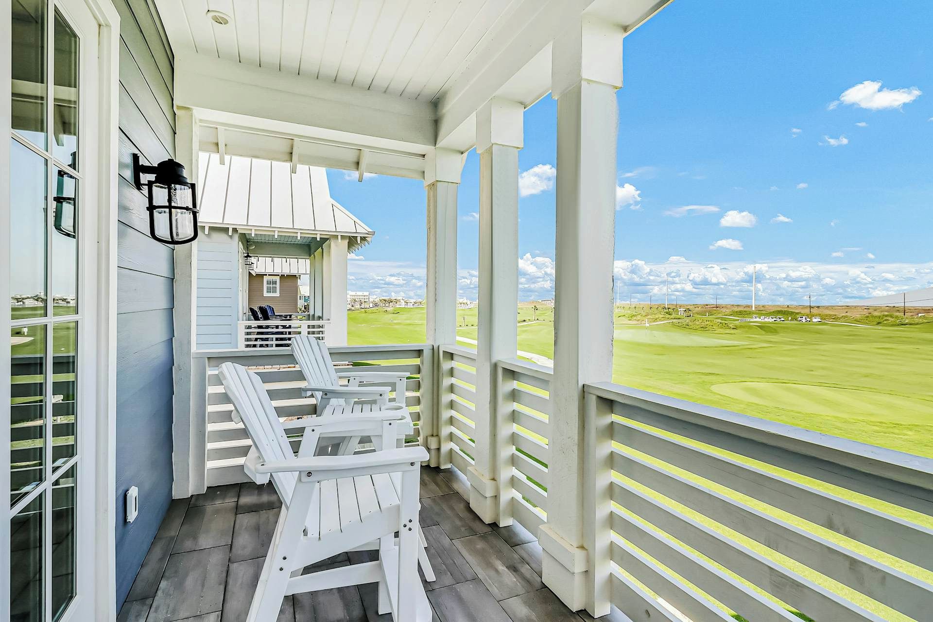 Eine überdachte Veranda mit weißen Adirondack-Stühlen, ein kleines Gebäude und Blick auf einen Golfplatz.