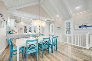 A spacious dining area with a white table and teal chairs, a white railing along the wall, and a large window with a view. - The Sand Trap | Pet Friendly | Steps to Beach (Port Aransas)