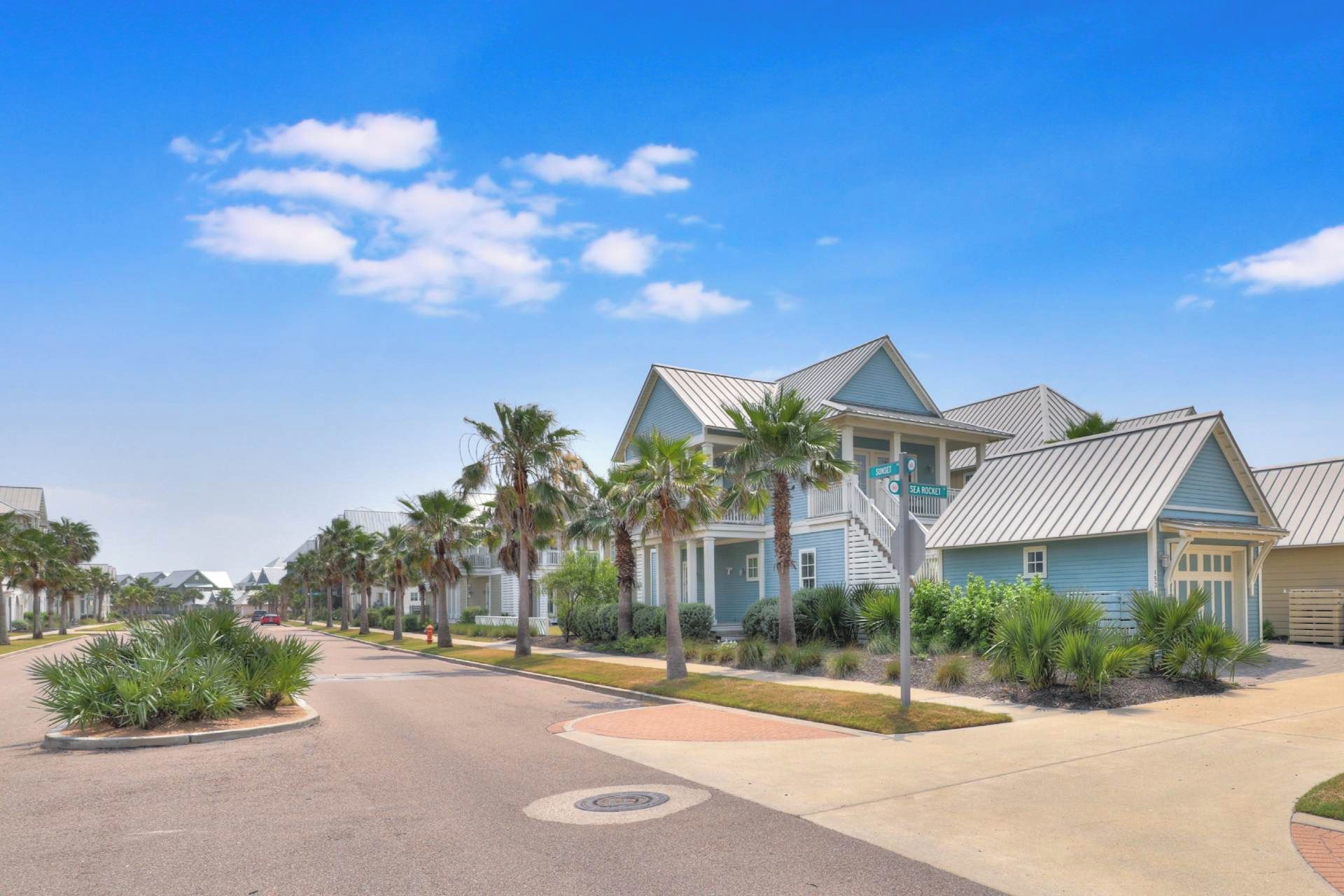 A coastal residential area with houses featuring blue exteriors and white railings, surrounded by palm trees and a clear sky.