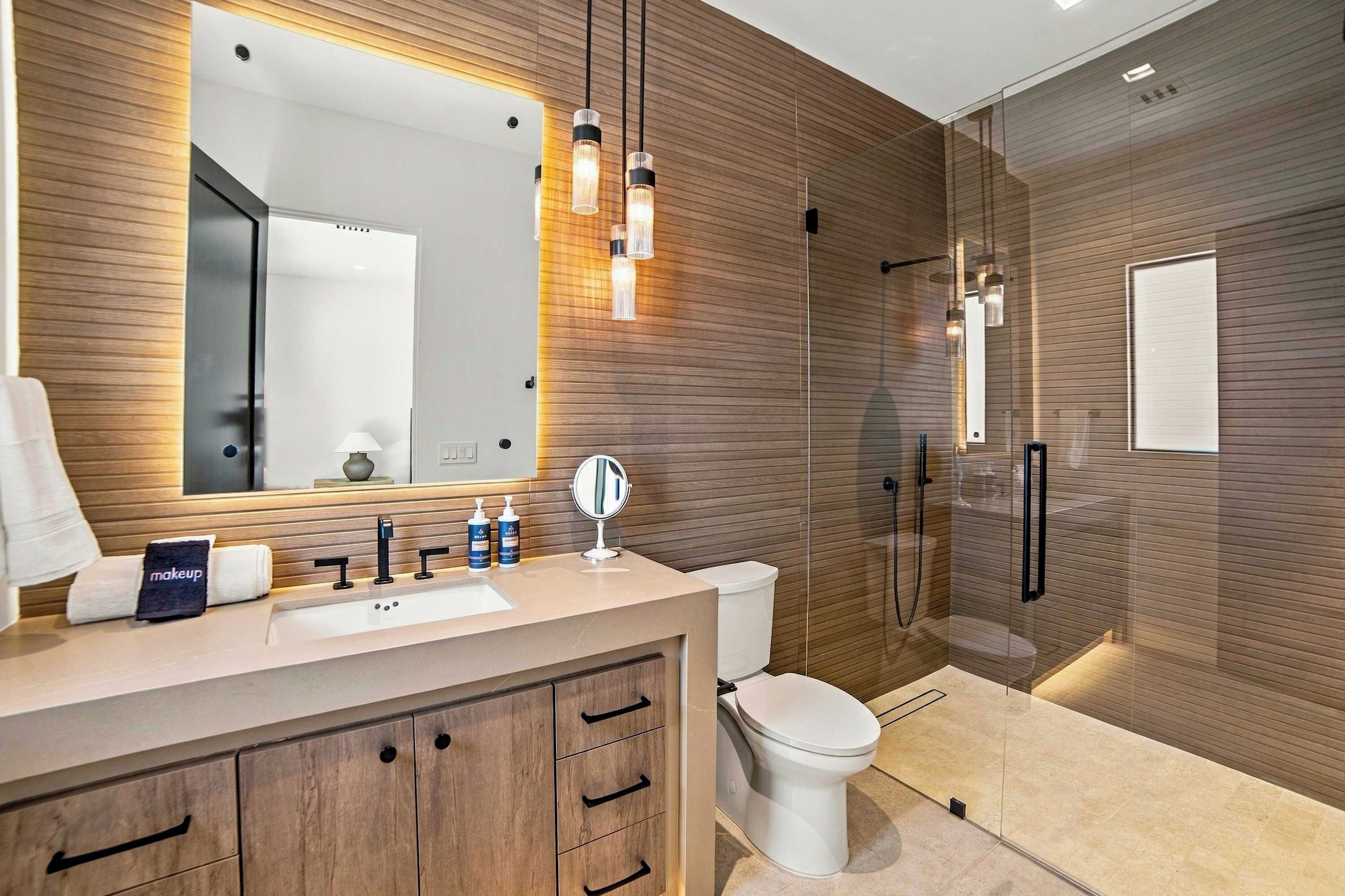 A modern bathroom with a wooden vanity, a large mirror, a toilet, and a glass-enclosed shower area.