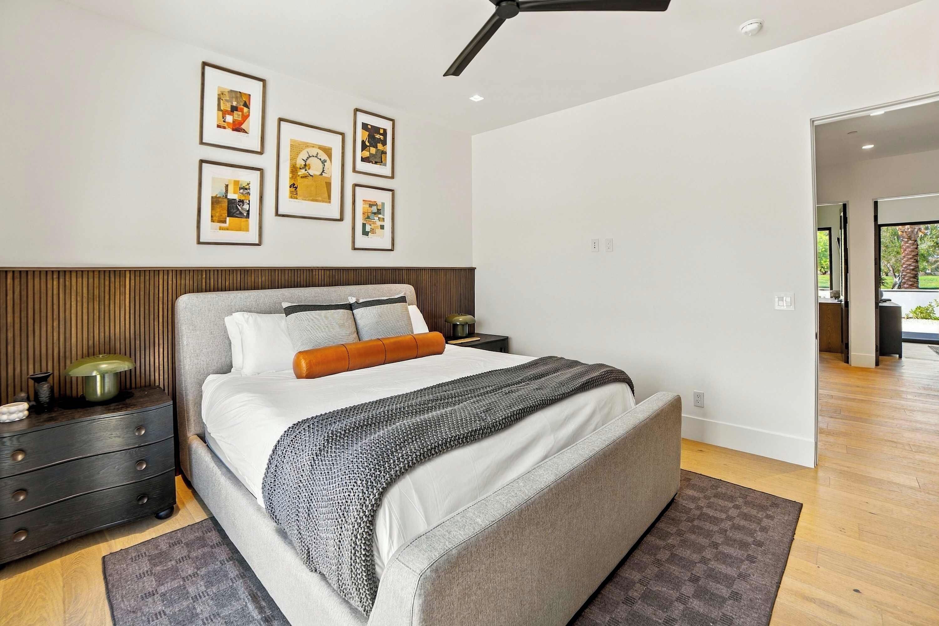 Une chambre à coucher moderne avec un lit, des tables de chevet, un ventilateur de plafond et des œuvres d’art encadrées accrochées au mur.