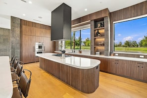A modern kitchen with a central island, built-in oven, and a large window offering a view of the outdoors. - HÓZHÓ| Golf Simulator, Theatre & Pickleball & Pool (Scottsdale)