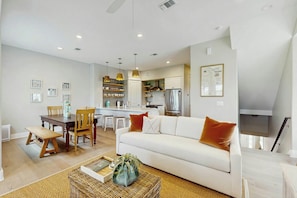 A modern living room with a white sofa, wooden dining table, and a kitchen in the background. - The Sandy Spur | Steps to Beach | Game Room (Port Aransas)