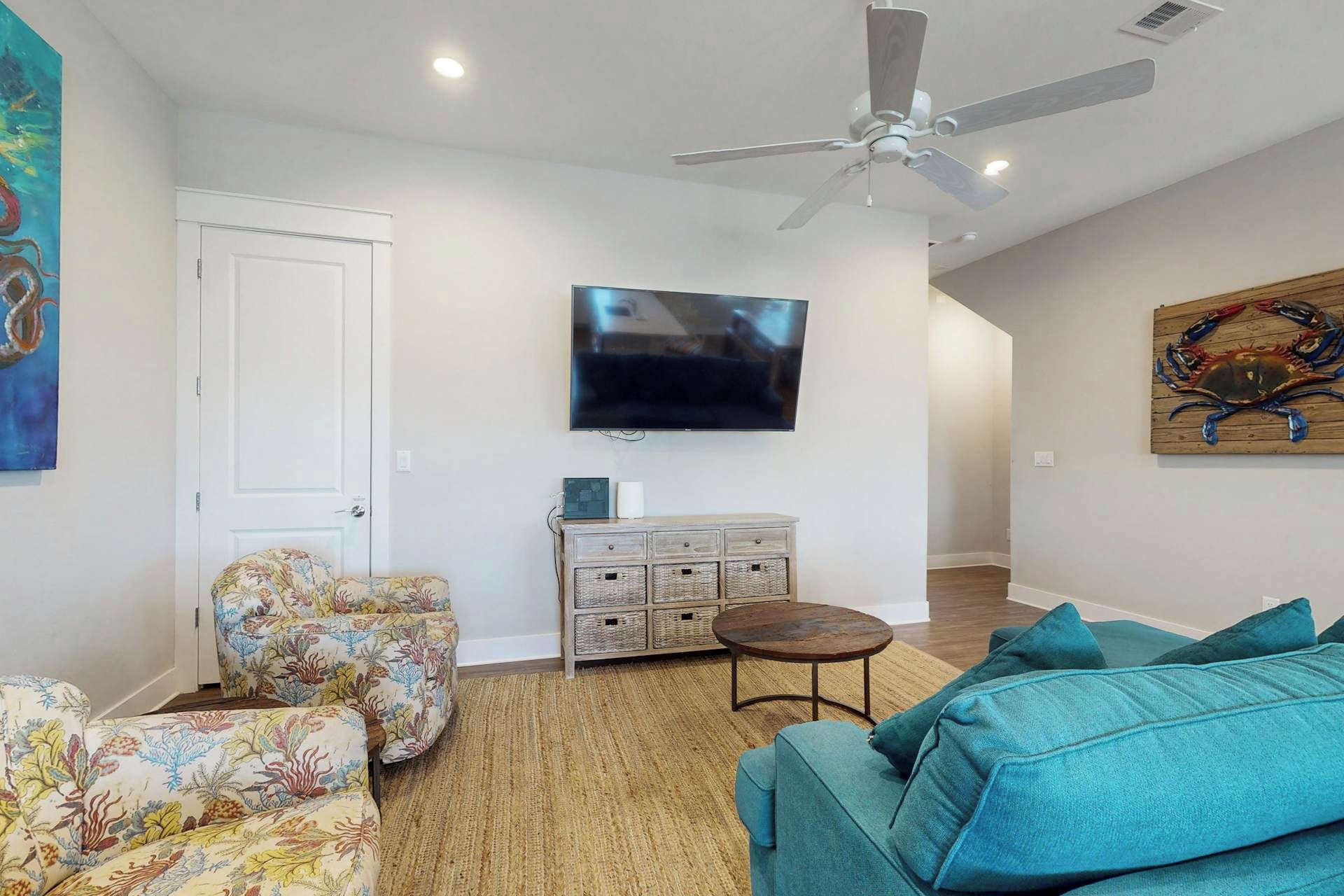 En stue med ventilator, fladskærms-tv, sofa, mønstret lænestol, lille bord og skab.