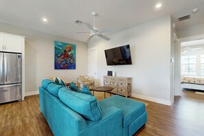 Ein modernes Wohnzimmer mit einem türkisfarbenen Sofa, einem Flachbildfernseher, einem Deckenventilator und einer Küchenzeile mit einem Edelstahlkühlschrank.