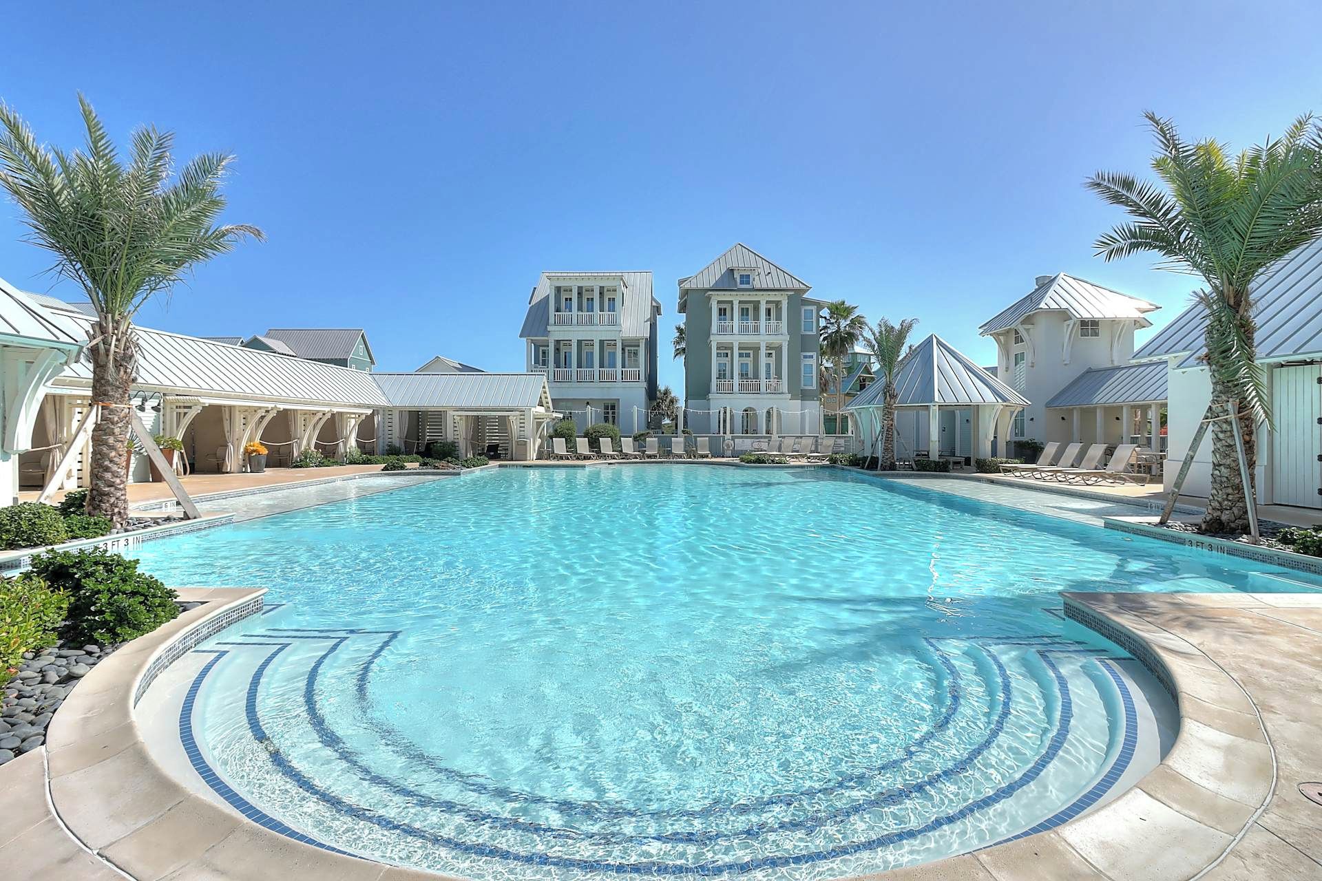 A large swimming pool with a clear blue center, surrounded by lounge chairs and palm trees, with a building in the background.