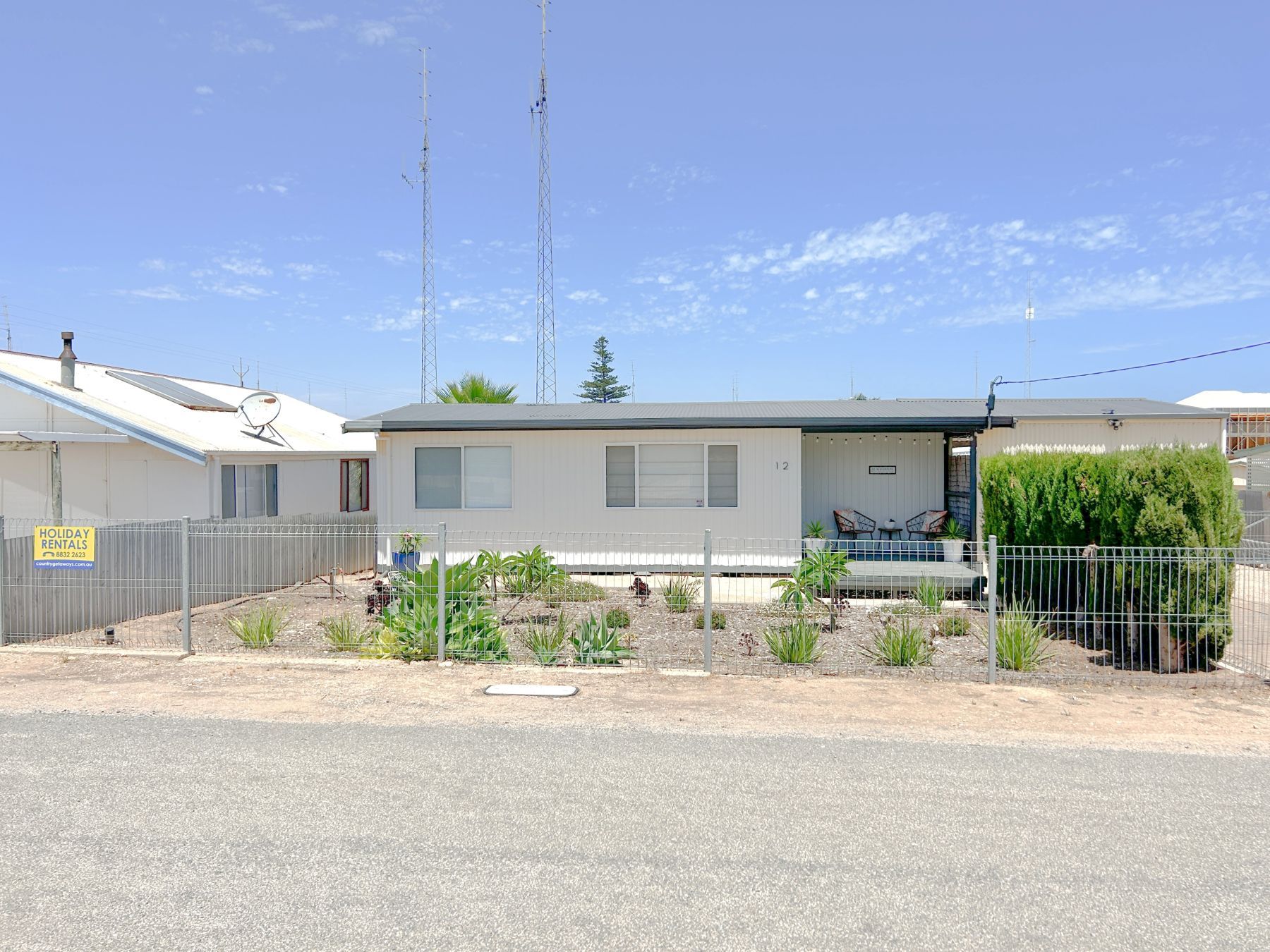 A single-story building with a fenced yard, a satellite dish, and a sign indicating holiday rentals.