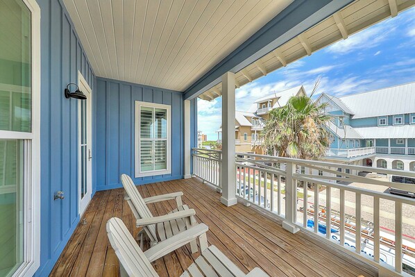 A covered porch with wooden flooring and white chairs, overlooking a coastal area with buildings and a palm tree. - Private Pool | Steps to Beach | Resort Amenities (Port Aransas)