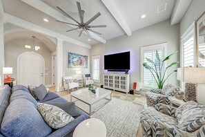 A spacious living room with a ceiling fan, a flat-screen TV, and a large window with blinds. - Private Pool | Steps to Beach | Resort Amenities (Port Aransas)