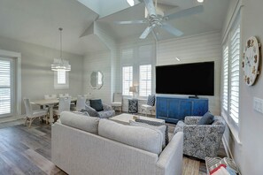 A spacious living room with a ceiling fan, a television, and a large window with blinds. - Green Turtle Cottage | Steps to Beach and Resort (Port Aransas)