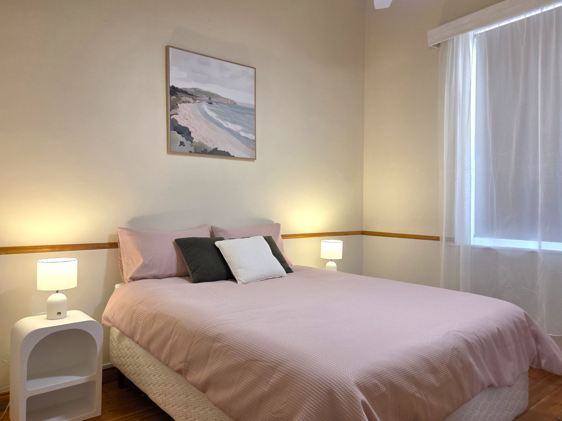 A bedroom with a bed, two bedside tables with lamps, a painting on the wall, and a window with curtains.