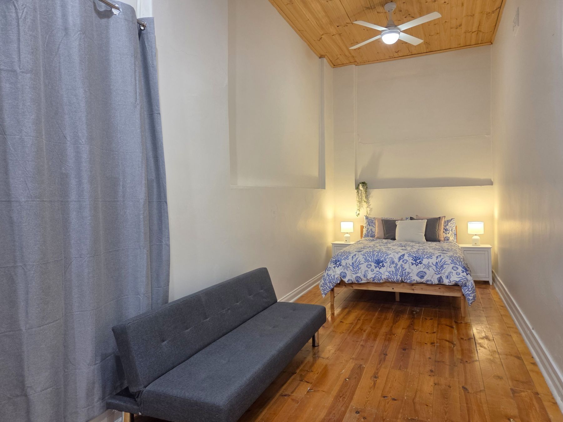A bedroom with a bed, bedside lamps, a ceiling fan, and a grey sofa.