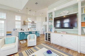 A modern kitchen with a large TV, white cabinetry, and a bar area with blue chairs. - Silverado Waves | Steps to Beach |Resort Amenities (Port Aransas)