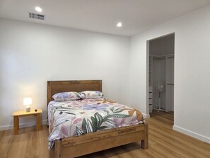 Un dormitori amb un llit de fusta, una tauleta de nit amb una làmpada i una paret blanca.