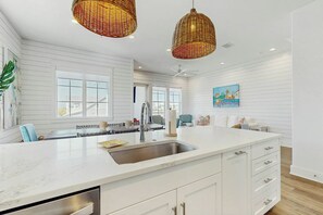 A modern kitchen with white cabinetry, a large island with a sink, and two wicker pendant lights. - Palmilla Beach CocoLoco| Gulf Views | Resort Perks (Port Aransas)