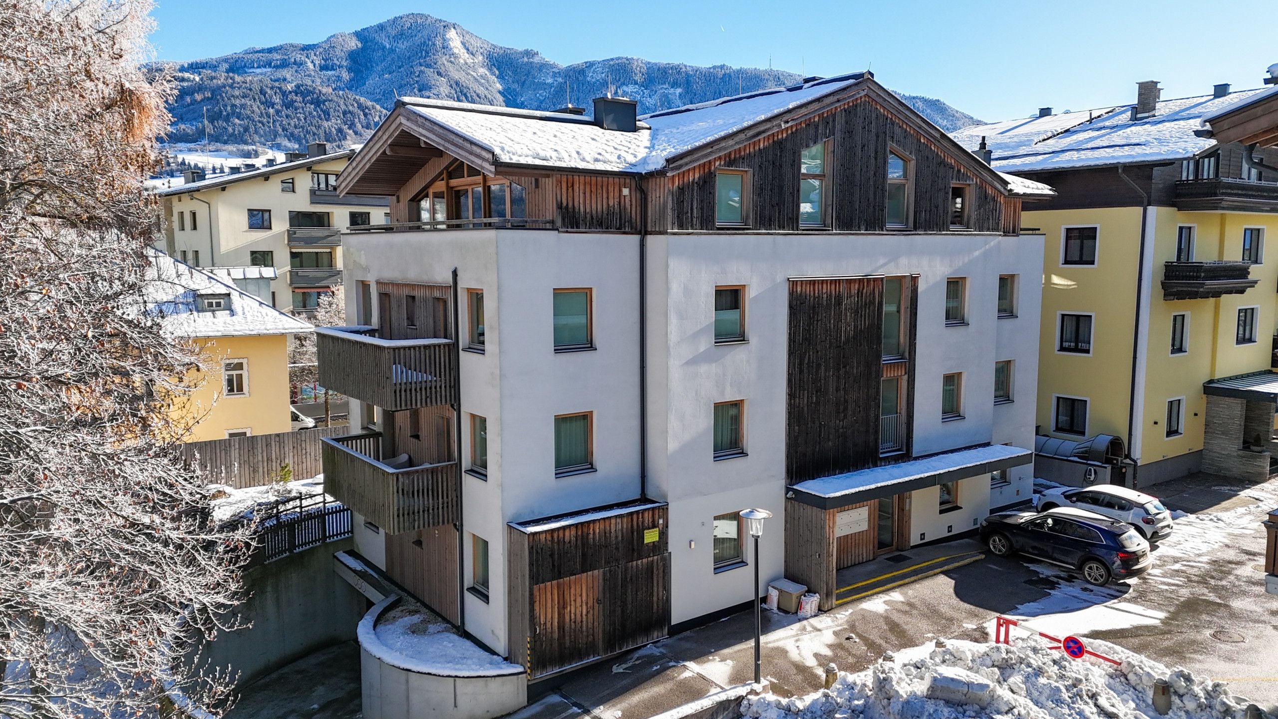A multi-story building with balconies, a parking area, and a snowy mountain backdrop.