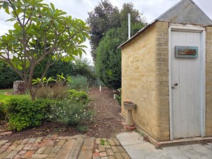 A small brick building with a door labeled 'TOILETTE' and a garden area with various plants and a tree.