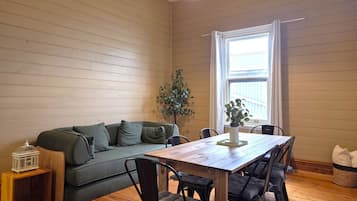 A cozy room with a wooden floor, a green sofa, a wooden dining table with chairs, a potted plant, and a window with curtains.