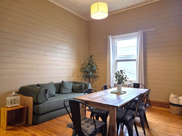 A cozy room with a wooden floor, a green sofa, a wooden dining table with chairs, a potted plant, and a window with curtains.