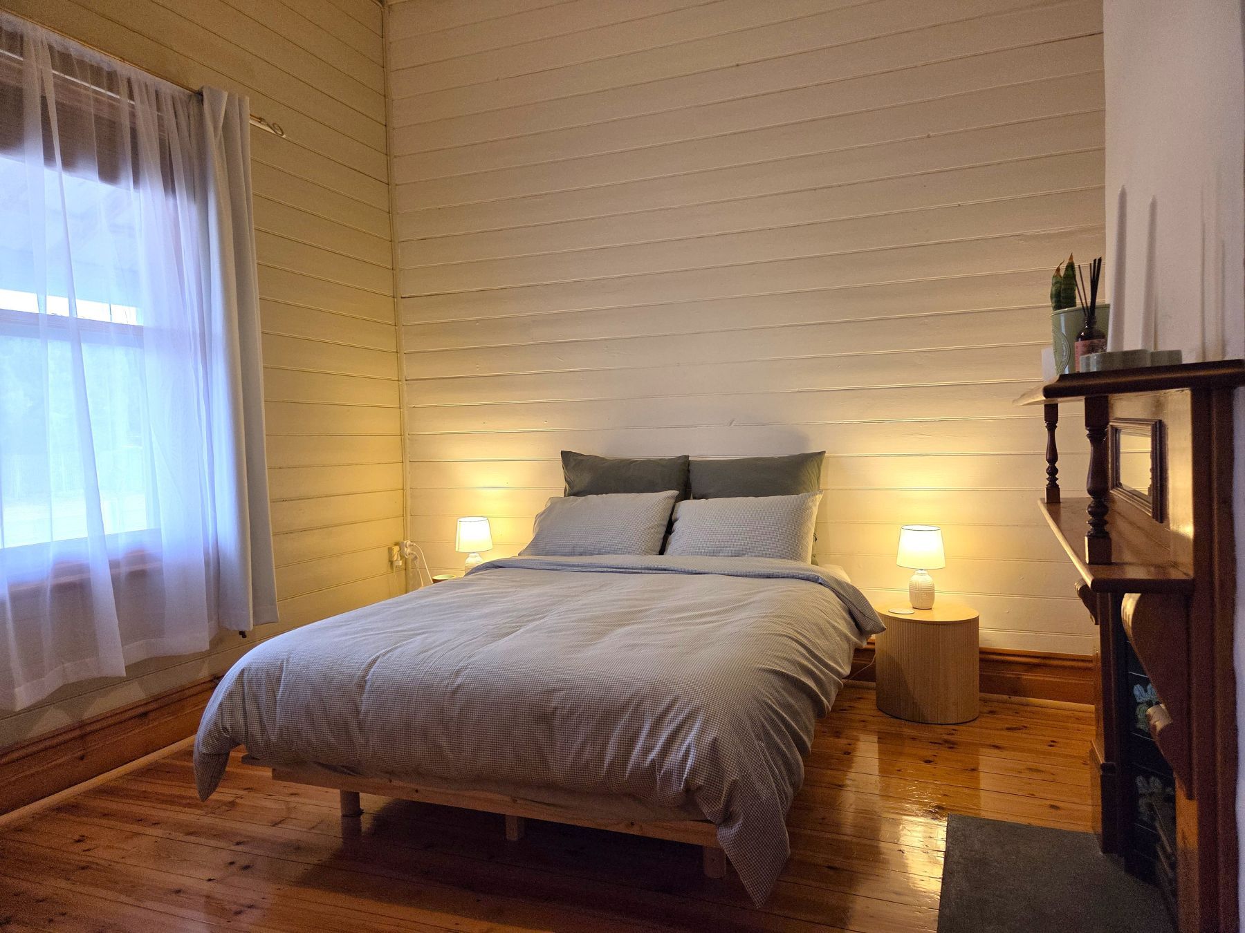 A bedroom with a wooden bed, two bedside lamps, a nightstand, and a window with sheer curtains.