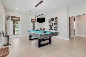 Een pooltafel in een kamer met een plafondventilator, een wandgemonteerde televisie en een deur die naar een andere kamer leidt.