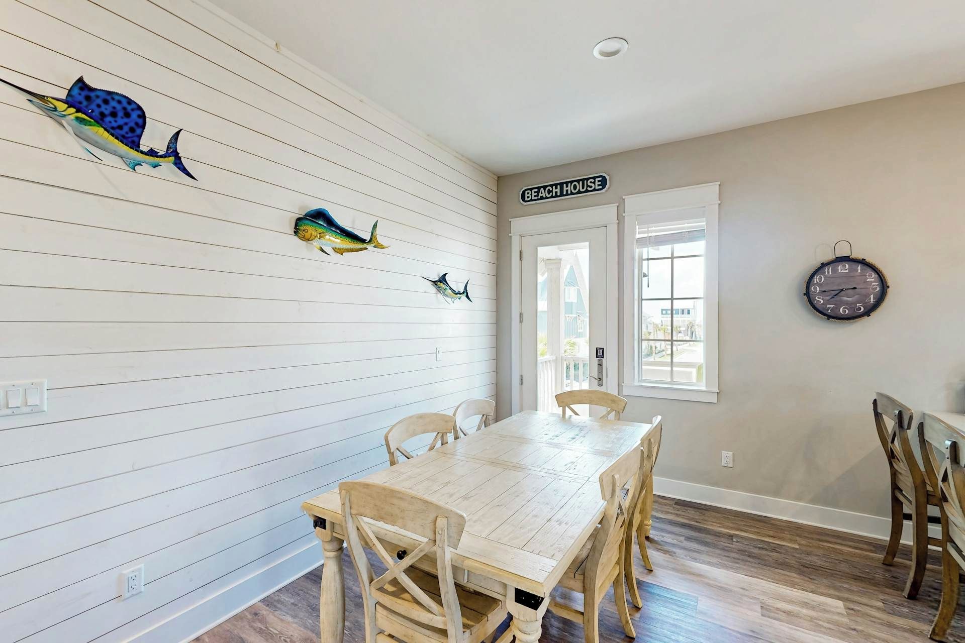 Een eetruimte met een houten tafel en stoelen, een klok aan de muur en een bordje met de tekst 'BEACH HOUSE'.
