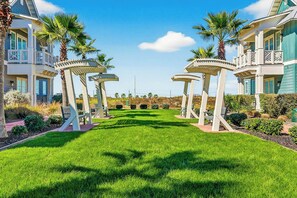 A well-maintained lawn with a gazebo structure, palm trees, and a clear sky. - Pines To Palms | Steps to Beach | Resort Amenities (Port Aransas)