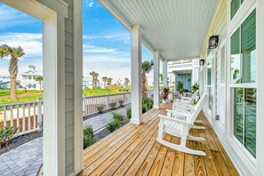A covered porch with wooden flooring, white rocking chairs, and a view of a residential area with houses and palm trees. - Salt and Serenity | Steps to Beach | Game Room (Port Aransas)