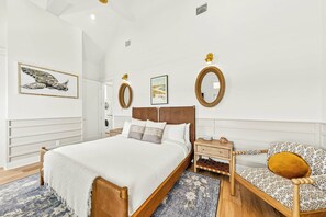 A bedroom with a large bed, a bedside table, a chair, and a wall-mounted mirror. - Salt and Serenity | Steps to Beach | Game Room (Port Aransas)