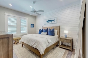 A bedroom with a bed, a nightstand, a lamp, a ceiling fan, a window with blinds, and a wall with a fish painting. - Sandy Fairway | Golf Views | Steps to Beach (Port Aransas)