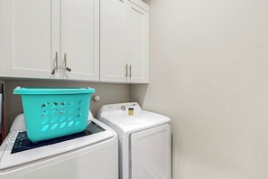 A laundry area with a washing machine and a dryer, a teal laundry basket on the machine, and white cabinets above. - Sandy Fairway | Golf Views | Steps to Beach (Port Aransas)