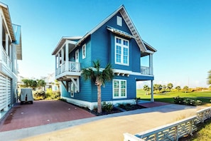 A two-story house with a balcony, a driveway, and a golf course in the background. - Sandy Toes | Golf Views | Pet Friendly (Port Aransas)