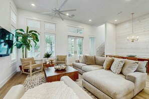 A spacious living room with a high ceiling fan, a large sofa, a coffee table, and a dining area with a chandelier. - Serenity Now | Gulf Views | Steps to Beach (Port Aransas)