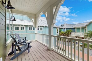A covered porch with two chairs and a table, overlooking a row of light blue buildings with white trim. - Serenity Now | Gulf Views | Steps to Beach (Port Aransas)