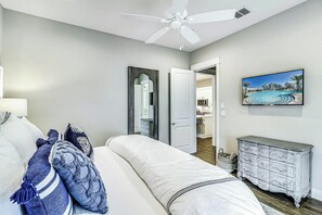 Une chambre à coucher comprenant un lit, un ventilateur de plafond, une commode, un tableau et une porte ouverte donnant sur une salle de bain.