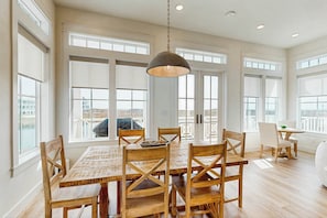 A spacious dining area with a large wooden table and chairs, large windows, and a hanging light fixture. - Songbird | Gulf Views | Private Elevator (Port Aransas)